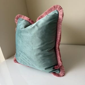 May include: A square throw cushion featuring a soft, sage green velvet fabric on the front and back. The edges of the cushion are finished with a decorative, dense fringe trim in a muted dusty pink colour that frames the entire perimeter.