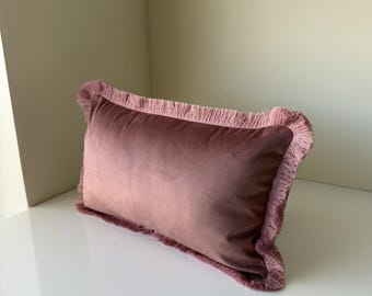 May include: A rectangular throw pillow in a dusty rose velvet fabric. The pillow features a matching fringe trim that runs along all four edges, creating a textured border around the smooth, soft-touch surface of the central velvet panel.