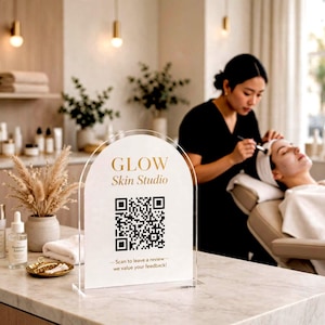 May include: An arched clear acrylic sign featuring the gold text GLOW Skin Studio, a black QR code, and the text Scan to leave a review we value your feedback. The sign is positioned on a light-coloured countertop in a professional setting.
