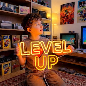 Pode incluir: Uma criança segura uma placa de néon laranja brilhante que diz "LEVEL UP". A placa apresenta um design de coração pixelado. O fundo inclui consoles de videogame retrô, caixas de jogos e pôsteres, criando um ambiente de jogo clássico.