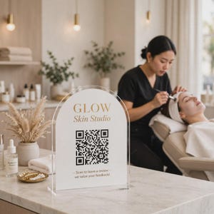 以下が含まれることがあります： 白い背景に金色の「GLOW Skin Studio」の文字、黒いQRコード、「Scan to leave a review we value your feedback」というテキストが印刷されたアーチ型のアクリル製サイン。透明な長方形のアクリルベースに取り付けられています。