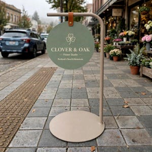May include: A circular sage green sign hanging from a beige metal stand with a round base. The sign features a white clover icon, the text CLOVER & OAK, Flower Studio, and Portland's Floral & Botanicals, attached by a brown leather strap.