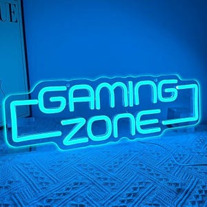 May include: A neon sign in the shape of the words "GAMING ZONE" in bright blue. The sign is illuminated against a blue background, with a white cable extending from the right side. The sign is placed on a textured surface.