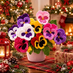 May include: A potted arrangement of crocheted pansy flowers in vibrant shades of purple, white, yellow, pink, and red, featuring green crocheted leaves. The flowers are displayed in a simple, solid light pink ceramic pot.