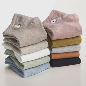 Könnte beinhalten: Stapel von Knöchelsocken in verschiedenen Farben, darunter Beige, Rosa und Grün. Einige Socken haben gestickte Tiermotive. Die Socken sind ordentlich gefaltet und gestapelt und zeigen eine Vielzahl von neutralen und pastellfarbenen Farbtönen.