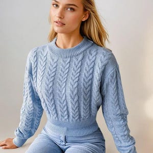 Puede incluir: Un suéter de punto trenzado azul claro con mangas largas y cuello redondo. El suéter se combina con vaqueros de mezclilla azul claro. La modelo tiene el pelo rubio.