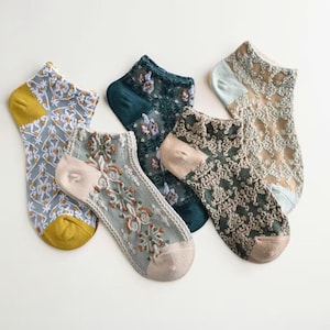 May include: An assortment of five ankle socks in various colors and patterns. The socks feature designs including floral embroidery, geometric shapes, and lace-like textures. Colors include gray, green, tan, and yellow.