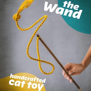 Può includere: Un giocattolo artigianale per gatti, con una bacchetta di legno con una corda e un nappa gialle. Il testo "the Wand" è visualizzato in bianco, con le parole "handcrafted cat toy" su uno striscione giallo.