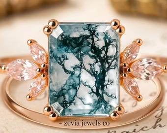 May include: Rose gold ring featuring a rectangular, light blue gemstone with a tree-like pattern. The ring has two clusters of pink, petal-shaped stones on either side of the main stone. The ring band is a delicate rose gold.