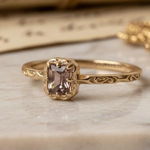 May include: A gold ring featuring a rectangular, pale brown gemstone. The ring has an ornate, floral design around the gemstone and on the band. The ring is set on a white marble surface, with a blurred background of a scroll and a gold chain.