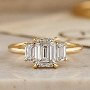 Op de afbeelding: Een gouden ring met drie diamanten in smaragdgroene slijpvorm. De middelste diamant is groter dan de twee zijstenen. De ring ligt op een licht oppervlak met een vage achtergrond.