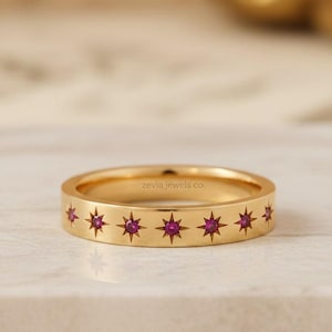 May include: A gold ring featuring star-shaped cutouts, each with a small, round, red gemstone. The ring is displayed on a neutral surface. The text "zevia jewels co." is visible on the ring. The ring's design is the focal point.