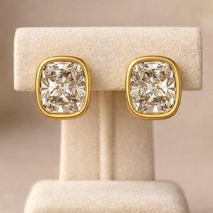 May include: A pair of gold-framed, square-cut stud earrings with a sparkling, clear center stone. The earrings are displayed on a beige jewelry stand, showcasing their elegant design and brilliance. The earrings are a classic jewelry item.