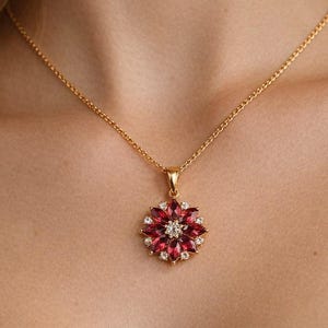 Op de afbeelding: Een goudkleurige ketting met een hanger in de vorm van een bloem. De hanger heeft rode en witte edelstenen. De ketting wordt om de nek gedragen.