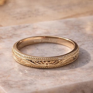 Könnte beinhalten: Ein goldfarbener Ring mit einem eingravierten floralen Muster und einem Perlenrand. Der Ring liegt auf einer Marmoroberfläche. Der Text "zevia jewels co" ist auf dem Ring sichtbar.
