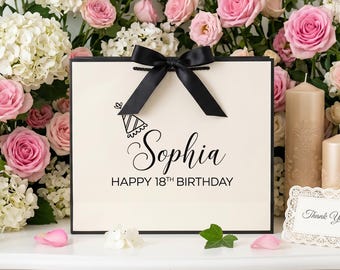 Pode incluir: Uma sacola de presente quadrada na cor creme com um laço de fita preta no topo. A frente apresenta um gráfico preto de um chapéu de festa, o nome Sophia em letra cursiva e o texto HAPPY 18TH BIRTHDAY em fonte sem serifa. Bordas pretas.