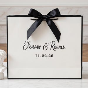 May include: A rectangular white gift bag with black trim and a large black satin ribbon bow at the top center. The front features the black printed text Eleanor & Rowan above the date 11.22.26 in a clean, bold, sans-serif font style.