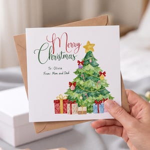 Puede incluir: Una tarjeta de felicitación cuadrada blanca con una ilustración en acuarela de un árbol de Navidad verde con una estrella amarilla, lazos rojos y adornos. El texto dice Merry Christmas, To: Olivia, From: Mom and Dad.
