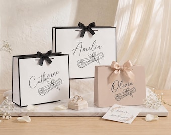 Op de afbeelding: Drie rechthoekige cadeauzakjes in wit en lichtroze, elk voorzien van een zwarte diploma-afbeelding met de tekst "GRADE 2027" en een gepersonaliseerde naam. De zakjes zijn afgewerkt met zwarte of champagnekleurige satijnen strikken.
