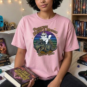 Könnte beinhalten: Hellrosa T-Shirt mit einem Aufdruck einer Katze mit Sonnenbrille und Krone, die auf einem Dinosaurier reitet. Der Text lautet "PRINCESS DONUT & MONGO" und "RIDE OR ROAR". Das Shirt ist ein Freizeitartikel.