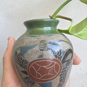 May include: A small, decorative vase with a turtle design. The vase features a brown turtle design on a blue background, with a green rim and a beige textured pattern. The vase is held in a hand.