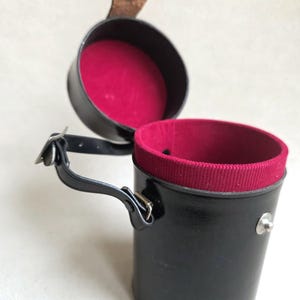 Può includere: Un astuccio cilindrico in pelle nera con interno in velluto rosso. L'astuccio ha un manico e un coperchio incernierato. Il coperchio è aperto, rivelando l'interno rosso. L'astuccio è probabilmente per riporre un piccolo oggetto.