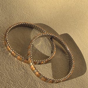 Peut inclure: Deux bracelets souples avec un motif répétitif de maillons rectangulaires argentés, dorés et or rose. Les bracelets sont circulaires et reposent sur une surface texturée brun clair, projetant une ombre.