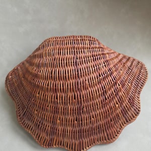 Könnte beinhalten: Ein geflochtener, brauner Weidenkorb in Muschelform. Der Korb hat einen gewellten Rand und eine strukturierte Oberfläche. Der Korb besteht aus dünnen, miteinander verwobenen Weidenstreifen.