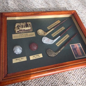 May include: A wooden-framed display case containing vintage golf memorabilia, including three miniature golf clubs, a silver-coloured woven ball, a red gutta-percha ball, a metal GOLF sign, and a small rectangular card featuring a golfer illustration.