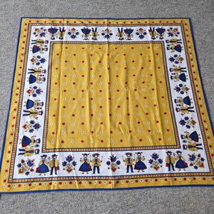Peut inclure: Une nappe carrée jaune avec une bordure bleue et une bordure à motifs blancs et jaunes. La nappe présente un motif répété de personnages en vêtements traditionnels et des accents floraux rouges et blancs.