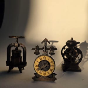 Op de afbeelding: Drie miniatuur metalen objecten in antieke stijl. De objecten omvatten een pers, een klok met Romeinse cijfers en een mechanisch apparaat met een wiel. De klok heeft een crèmekleurige wijzerplaat en zwarte wijzers.