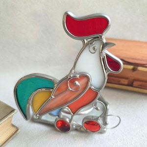 Puede incluir: Figura de gallo de vidriera colorida, con cresta roja, cuerpo naranja y blanco, y cola verde. El gallo tiene un marco de metal plateado y pies de cristal rojo. Artículo decorativo para el hogar.