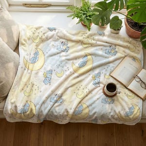 Peut inclure: Une couverture douce de couleur claire avec un motif répétitif d'oursons bleus dessinés se reposant sur des croissants de lune jaunes entourés de petites étoiles. La couverture est étalée sur un banc de fenêtre près de deux coussins beiges.