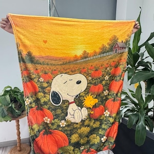 Peut inclure: Une couverture rectangulaire en polaire illustrant une scène de champ de citrouilles en automne. Snoopy et Woodstock sont au centre parmi des citrouilles orange et du feuillage vert. Le fond montre un coucher de soleil doré et une grange rouge.