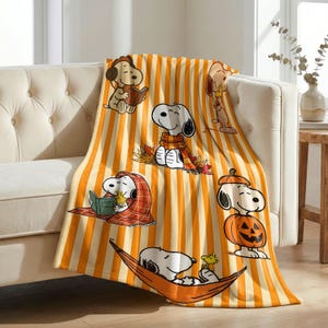 Könnte beinhalten: Eine weiche Wohndecke mit einem vertikalen Streifenmuster in Orange und Creme. Die Decke zeigt verschiedene Illustrationen von Snoopy und Woodstock, darunter Snoopy beim Lesen eines Buches, mit einem Schal, in einem Kürbis sitzend und in einer Hängematte ruhend, alles vor dem gestreiften Hintergrund.