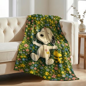 Op de afbeelding: Een zachte fleece deken met een groene achtergrond bedekt met een dicht patroon van kleine roze, gele en blauwe bloemen. Het midden toont een grote illustratie van Snoopy en Woodstock, waarbij Snoopy kleine bloemaccenten draagt.