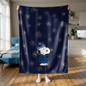 Pode incluir: Uma manta de lã azul-marinho escuro com um padrão repetitivo de flocos de neve brancos e um gráfico central da personagem Snoopy a usar um gorro de inverno azul e um casaco acolchoado a condizer, representado de perfil.