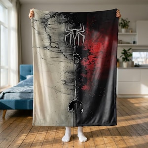 Può includere: Una coperta rettangolare con un design diviso, caratterizzata da un lato color crema con texture nere screpolate e un lato rosso intenso e nero. Un motivo centrale raffigura un emblema di ragno nero e una figura sagomata appesa a una ragnatela.