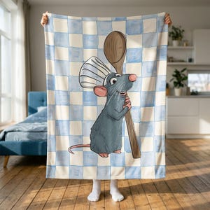 Peut inclure: Une couverture en polaire douce représentant un rat de dessin animé portant une toque de chef blanche et tenant une grande cuillère en bois. Le fond présente un motif à carreaux bleu clair et crème. Le rat a un pelage gris et des oreilles roses.