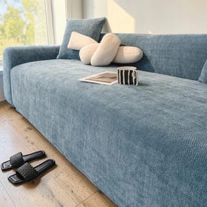 May include: A light blue sofa with a textured herringbone pattern. Decorative cushions in light blue and cream colours rest on the sofa. A black and white striped mug and a pair of black woven sandals are also visible.