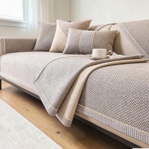 May include: A beige sofa with a woven fabric cover and matching throw pillows. The sofa has a neutral colour palette with a light beige base and darker beige accents. A white teacup sits on a small tray on the sofa.