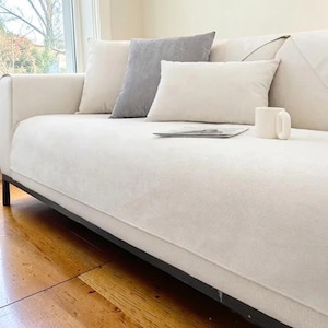 以下が含まれることがあります： 白とグレーのクッションがいくつか置かれた、黒い金属フレームのクリーム色のソファ。白いマグカップと雑誌がソファの上にあります。ソファは窓の前に配置されています。