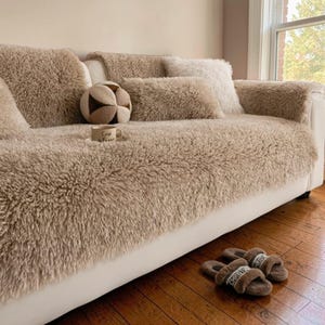 May include: A beige, fluffy sofa cover and matching cushions are arranged on a white sofa. A decorative ball, a coffee cup, and a pair of brown slippers are on the sofa. The slippers have the word "CHRISTIAN" on them.