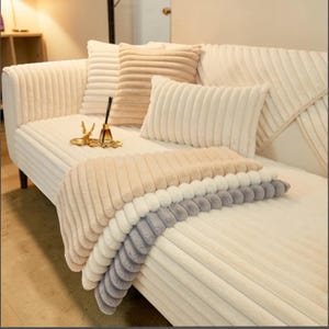 Puede incluir: Un sofá color crema con cojines a juego y una manta plegada. Los cojines y la manta tienen una textura acanalada, con la manta con capas de beige, blanco y gris. Objetos decorativos están en el sofá.