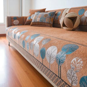 May include: A sofa with an orange cover displaying a pattern of blue, white, and grey stylized trees. Matching cushions and a decorative ball are also visible. The sofa has a light-coloured frame and is placed on wooden flooring. The sofa's dimensions are in centimetres.