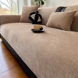 Puede incluir: Un sofá beige con dos cojines a juego y una pelota con estampado en blanco y negro. Una taza pequeña y un platillo están en el sofá. El sofá tiene una base y reposabrazos negros.