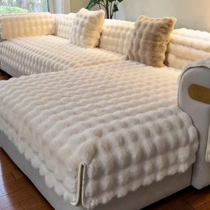 May include: A sectional sofa with a cream-colored, textured, faux fur cover. The sofa has several matching throw pillows in varying sizes and shades of cream and beige. The fabric has a soft, plush appearance, creating a cozy aesthetic.