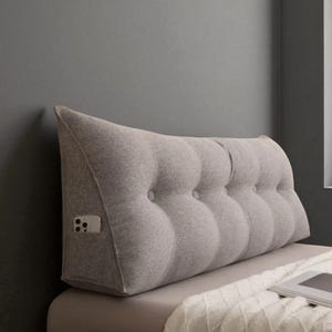May include: A grey, wedge-shaped bed rest pillow with a button-tufted design. The pillow has a pocket on the side for a phone. It is placed on a bed with a white blanket.