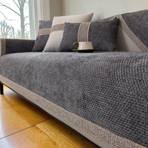 May include: A gray sofa with matching throw pillows and a textured fabric cover. The pillows have neutral-toned accents. A cup and saucer sit on the sofa. The sofa is placed in front of a window with natural light.