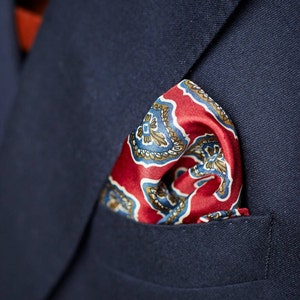 May include: A red and blue patterned pocket square with a paisley design. The pocket square is folded and placed in the pocket of a navy blue blazer.